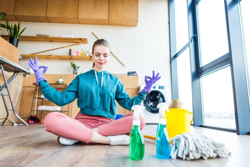 How to Keep Your House Clean When You are Away smiling young woman sitting on floor and meditating while cleaning house e1669624929148