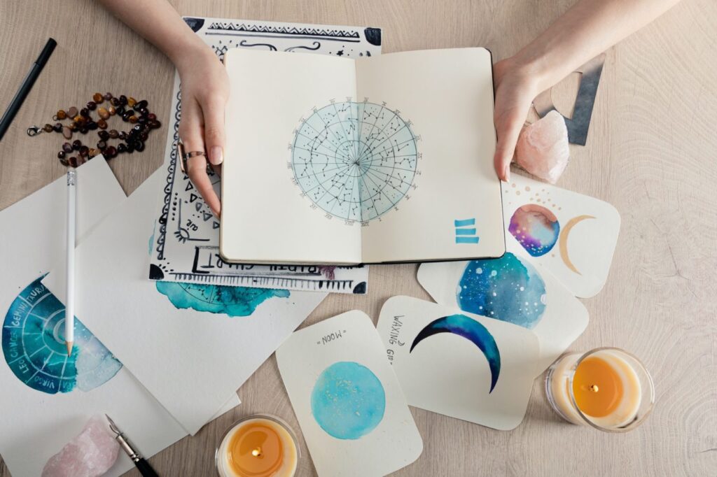 Find Your Best House Using Astrology & Sign top view of astrologer holding notebook with watercolor drawings and zodiac signs on cards on table e1669624941145